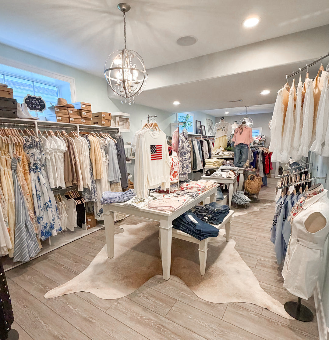 A boutique clothing store interior with racks of dresses, shirts, and folded garments, a central white display table with folded jeans and a shirt featuring an American flag graphic, a white animal-print rug on the wood floor, and a hanging globe chandelier.